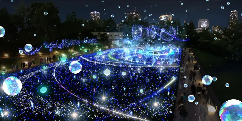ภาพสวนสาธารณะในเวลากลางคืนที่ประดับด้วยไฟสีน้ำเงินและวงกลมเรืองแสงจำนวนมาก มีผู้คนเดินอยู่ตามทางเดินและอาคารสูงอยู่เบื้องหลัง