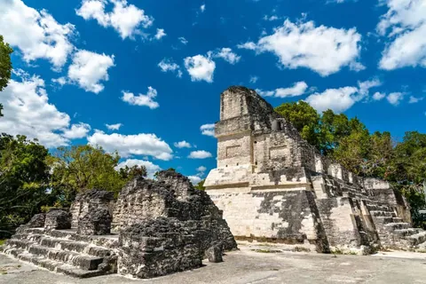 โครงสร้างโบราณสถานหินสีขาวมีบันไดและยอดสูงตั้งอยู่ท่ามกลางต้นไม้เขียวขจี ใต้ท้องฟ้าสีน้ำเงินเข้มที่มีเมฆสีขาวลอยอยู่