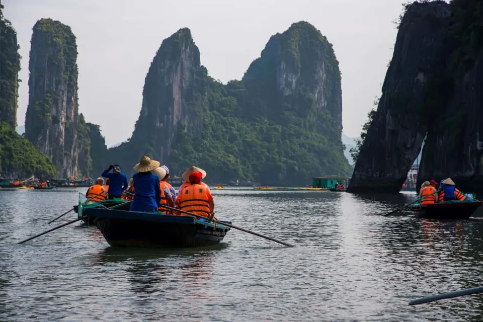 เรือไม้ลำเล็กที่มีผู้โดยสารสวมเสื้อชูชีพสีส้มและหมวกฟางกำลังแล่นอยู่บนผิวน้ำ โดยมีหน้าผาหินสูงชันปกคลุมด้วยพืชสีเขียวตั้งตระหง่านอยู่เบื้องหลัง