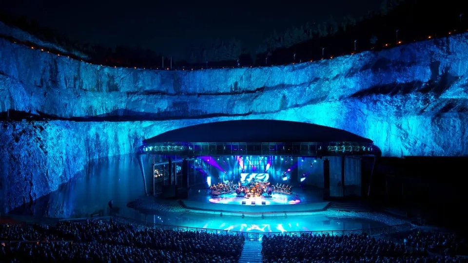 ภาพเวทีแสดงกลางแจ้งขนาดใหญ่ตั้งอยู่ภายในแอ่งหินที่ถูกส่องด้วยแสงสีน้ำเงิน มีผู้ชมจำนวนมากนั่งอยู่รอบๆ