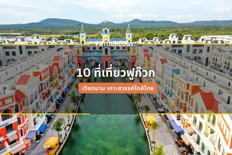 ภาพมุมสูงของคลองสีเขียวขุ่นที่ไหลผ่านระหว่างอาคารหลายชั้นสีสดใส มีสะพานสีน้ำเงินและส้มทอดข้ามคลอง โดยมีภูเขาเป็นฉากหลัง