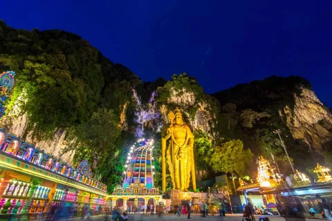 รูปปั้นสีทองขนาดใหญ่ตั้งอยู่หน้าหน้าผาหินที่มีต้นไม้ปกคลุมในเวลากลางคืน โดยมีอาคารประดับไฟหลากสีสันอยู่ด้านซ้ายและผู้คนเดินอยู่บริเวณด้านล่าง
