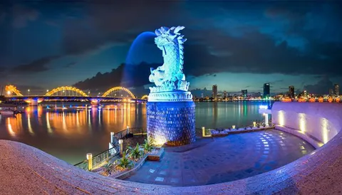 รูปปั้นมังกรสีฟ้าขนาดใหญ่ตั้งอยู่บนฐานกลมริมแม่น้ำในเวลากลางคืน โดยมีสะพานโค้งหลายช่วงและแสงไฟเมืองสะท้อนอยู่บนผิวน้ำเบื้องหลัง