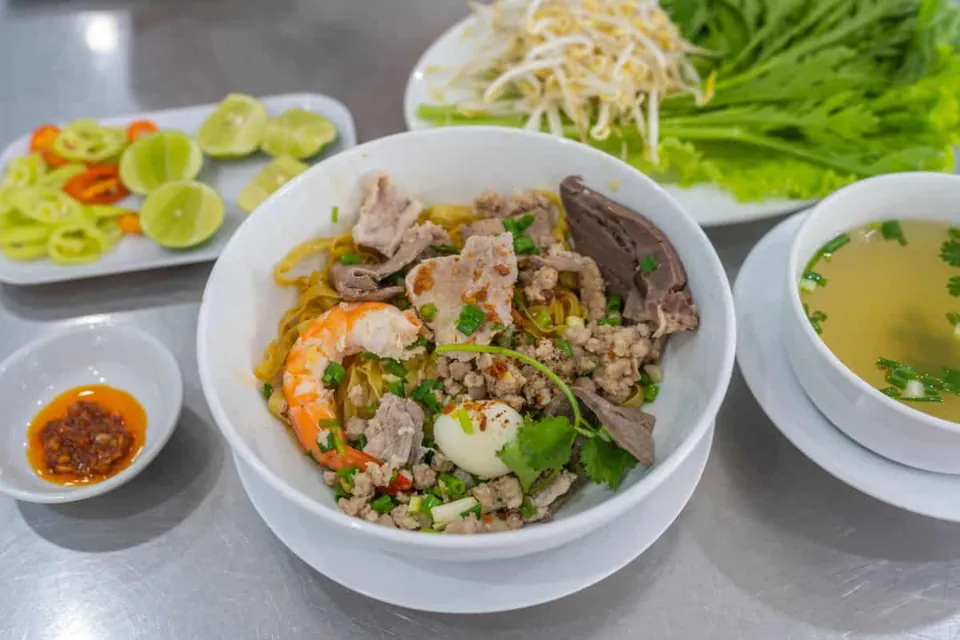 ชามข้าวซอยพร้อมกุ้ง ไข่ต้ม และเนื้อสัตว์ วางบนโต๊ะโลหะ มีชามซุป ผักสด และน้ำจิ้มสีแดงข้างเคียง
