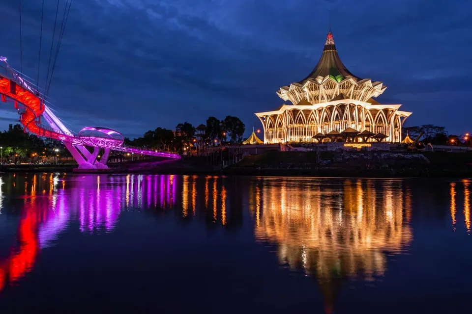 ภาพอาคารทรงหลังคาแหลมประดับไฟสีเหลืองทองตั้งอยู่ริมแม่น้ำในเวลากลางคืน มีสะพานรูปทรงโค้งประดับไฟสีแดงและสีม่วงอยู่ด้านซ้าย สะท้อนลงบนผิวน้ำ