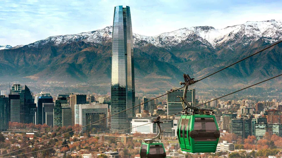 กระเช้าสีเขียวสองคันแขวนอยู่บนสายเคเบิลเหนือเมืองที่มีตึกสูงและภูเขาหิมะเป็นฉากหลัง