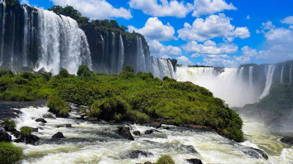 ภาพน้ำตกขนาดใหญ่หลายสายไหลลงมาจากหน้าผาหินสีดำ มีพุ่มไม้สีเขียวปกคลุมเกาะกลางน้ำและขอบหน้าผา ใต้ท้องฟ้าสีฟ้าที่มีเมฆสีขาวลอยอยู่
