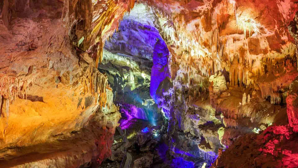 ภาพภายในถ้ำที่มีหินงอกหินย้อยเป็นชั้นๆ สว่างไสวด้วยแสงไฟสีส้ม แดง และม่วงที่ส่องกระทบพื้นผิวหิน