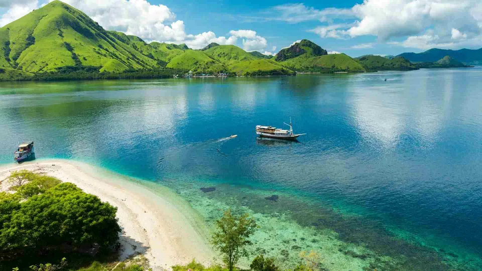 ภาพมุมสูงของทะเลสีฟ้าครามที่มีเรือสองลำลอยอยู่กลางน้ำ ข้างๆ เป็นหาดทรายสีขาวและภูเขาเขียวขจี