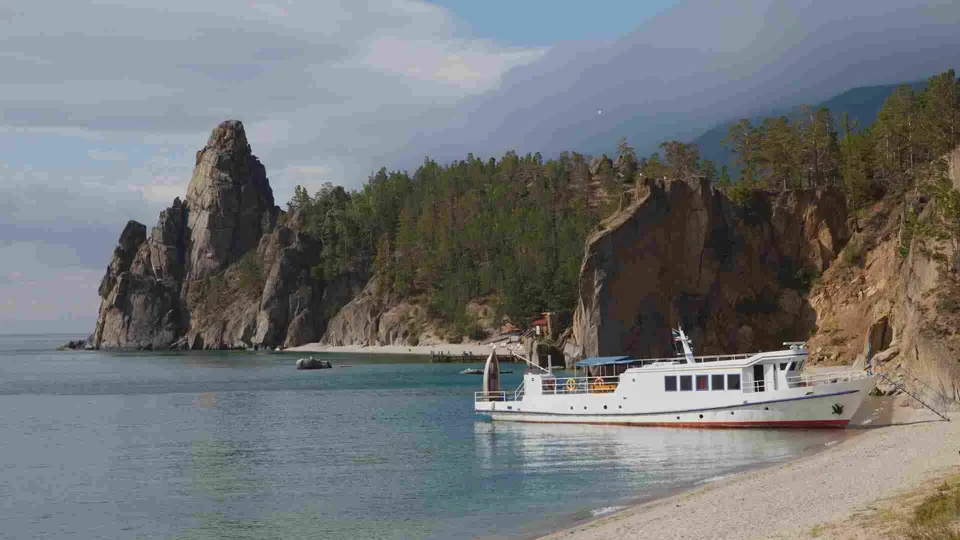 เรือสีขาวลำหนึ่งจอดอยู่ริมชายหาดหินกรวดใกล้หน้าผาหินสูงที่ปกคลุมด้วยต้นไม้สีเขียวริมทะเล