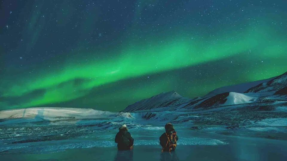 ภาพทิวทัศน์ยามค่ำคืนที่มีแสงเหนือสีเขียวเรืองรองบนท้องฟ้าเหนือภูมิประเทศหิมะและภูเขา โดยมีบุคคลสองคนยืนอยู่บริเวณพื้นน้ำแข็งด้านหน้า
