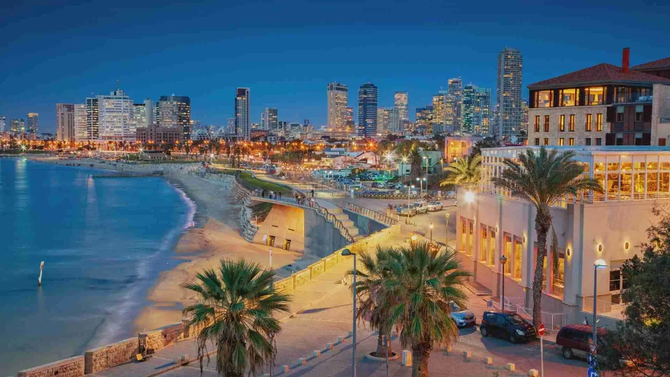 ภาพมุมสูงของชายหาด Tel Aviv ในยามค่ำ มีถนนริมทะเล ปาล์ม และอาคารสูงเรียงรายอยู่เบื้องหลัง