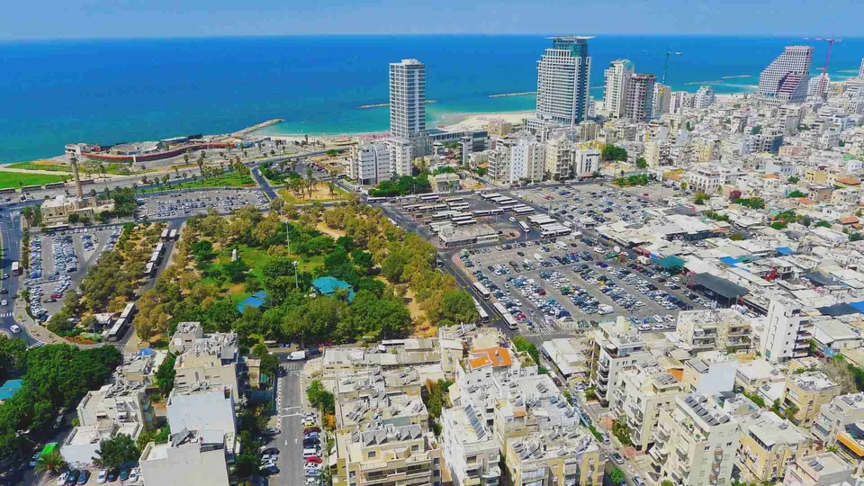 ภาพมุมสูงแสดงเมือง Tel Aviv ติดกับทะเลสีฟ้า มีอาคารสูงหลายหลังตั้งอยู่ริมชายหาด และลานจอดรถขนาดใหญ่ในบริเวณใกล้เคียง