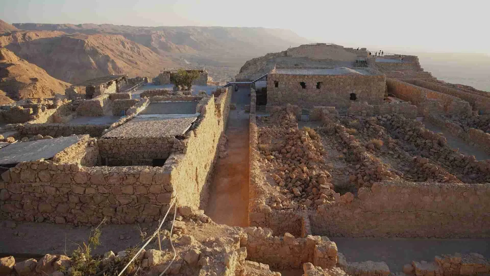 ภาพซากปรักหักพังของโครงสร้างหินบนยอดเขา Masada โดยมีเนินเขาและทะเลสาบอยู่เบื้องหลัง