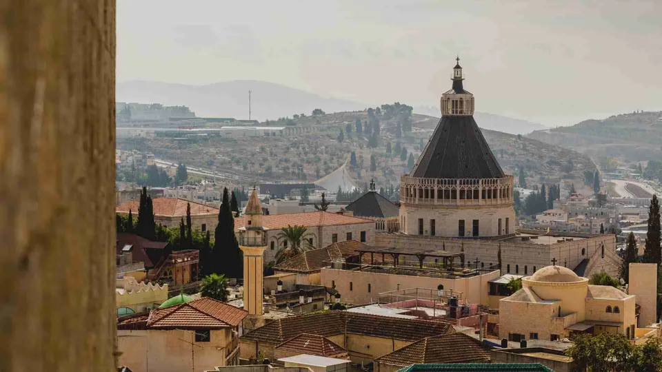 ภาพทิวทัศน์เมือง Nazareth แสดงอาคารโดมสีดำขนาดใหญ่ตั้งอยู่ท่ามกลางบ้านเรือนหลังเล็กและเนินเขาในพื้นหลัง