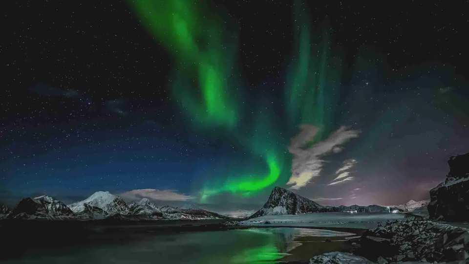 ภาพทิวทัศน์ยามค่ำคืนที่มีแสงสีเขียวเรืองรองเป็นริ้วในท้องฟ้าเหนือยอดเขาและผิวน้ำที่สะท้อนแสง