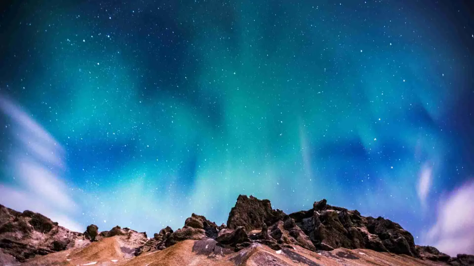 ภาพทิวทัศน์ยามค่ำคืนที่มีแสงสีเขียวอมฟ้าเรืองรองเป็นริ้วในท้องฟ้าเหนือแนวหินและเนินดินสีน้ำตาล