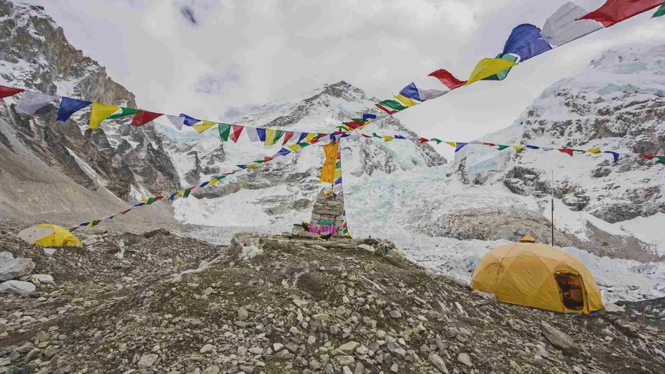 ภาพทิวทัศน์ภูเขาหิมะที่มีเต็นท์สีเหลืองตั้งอยู่บนพื้นหินและสายธงสีรุ้งพาดผ่านกลางภาพ