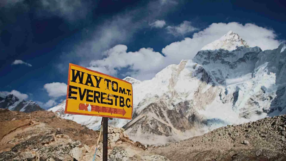 ป้ายสีเหลืองเขียนว่า 'WAY TO M.T. EVEREST B.C.' ตั้งอยู่หน้าภูเขาสูงปกคลุมด้วยหิมะและก้อนหิน