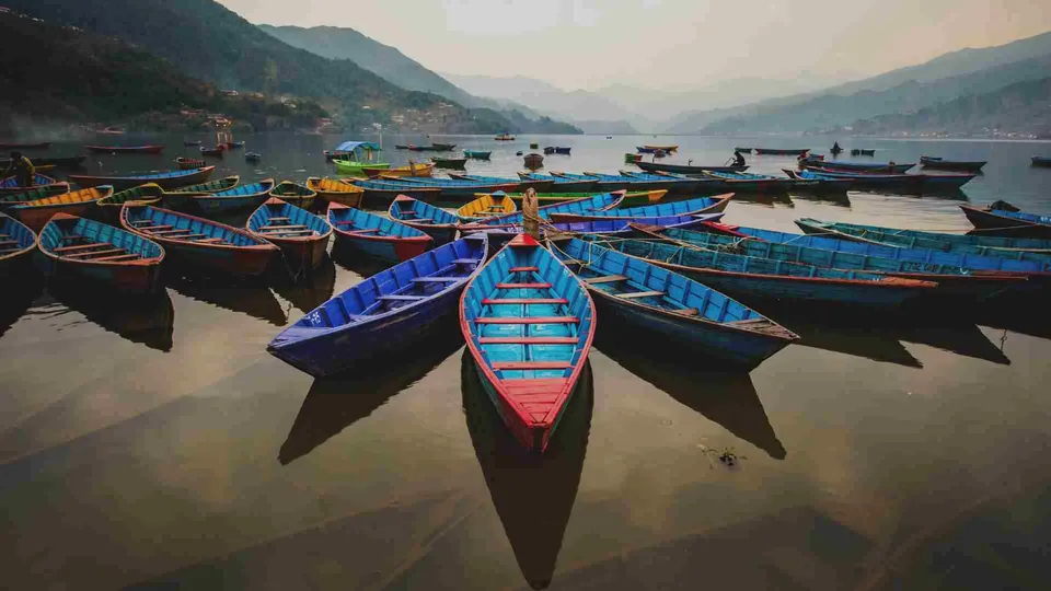 เรือไม้หลายลำที่ทาสีน้ำเงินและแดงจอดเรียงกันบนผิวน้ำที่สงบ โดยมีทิวเขาสีเขียวทอดตัวเป็นฉากหลัง