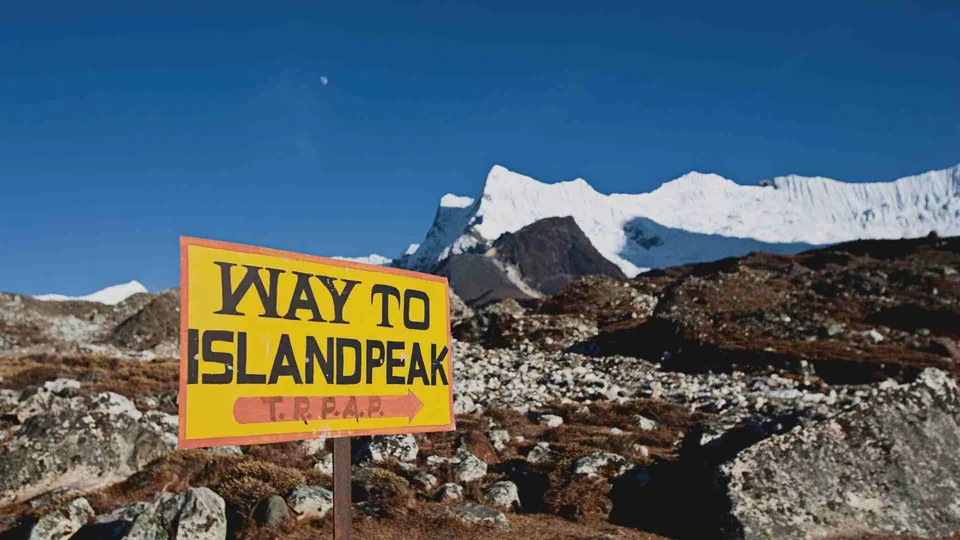 ป้ายสีเหลืองข้อความ 'WAY TO ISLANDPEAK' ตั้งอยู่บนพื้นหินและดินสีน้ำตาล โดยมีภูเขาปกคลุมด้วยหิมะเป็นฉากหลังภายใต้ท้องฟ้าสีน้ำเงิน