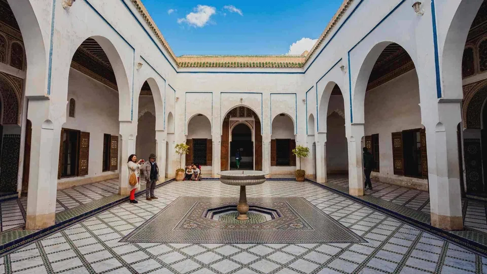 ลานcourtyardสีขาวที่มีซุ้มประตูโค้งเรียงราย พื้นปูด้วยกระเบื้องลายตารางสีน้ำเงินและขาว มีอ่างน้ำพุตั้งอยู่ตรงกลาง และมีผู้คนยืนอยู่บริเวณด้านซ้าย