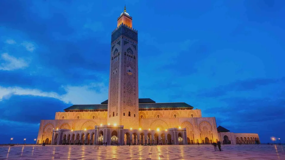 มัสยิดฮัสซันที่ 2 (Hassan II Mosque) สถาปัตยกรรมสีขาวขนาดใหญ่พร้อมหอคอยสูง ตั้งอยู่บนลานกว้างที่สะท้อนแสงไฟสีเหลืองอุ่น ภายใต้ท้องฟ้าสีน้ำเงินเข้ม
