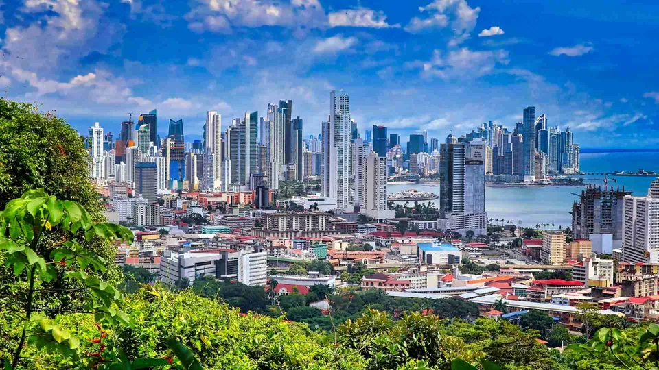 ภาพทิวทัศน์เมืองที่มีอาคารสูงเรียงรายติดชายฝั่งทะเล มีกลุ่มอาคารสูงเด่นอยู่ตรงกลาง และพุ่มไม้สีเขียวอยู่ด้านหน้า