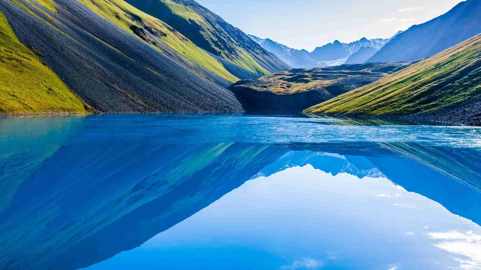 ภาพทิวทัศน์ของทะเลสาบสีน้ำเงินเข้มที่สะท้อนเงาของภูเขาสูงชันปกคลุมด้วยหญ้าสีเขียวและหินสีเทาในหุบเขา
